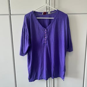 May include: A vibrant purple short-sleeved shirt featuring a vertical ribbed texture, a V-neckline, and a partial button-down placket with five small buttons centered on the chest. The garment has a relaxed fit and a straight hemline with side slits.