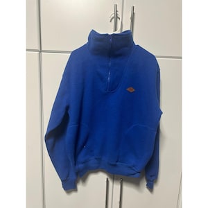 Op de afbeelding: Een levendige koningsblauwe fleece trui met een hoge kraag en een kwartlange ritssluiting. Het kledingstuk is voorzien van een kleine, ruitvormige bruine leren patch op de linkerborst en geribbelde boorden aan de mouwen en de onderkant.