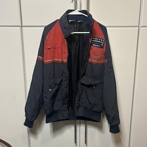 May include: A navy blue and red jacket with a high collar, front zipper, and ribbed cuffs. The chest features a rectangular patch with the text UNITED AVIATION ELECTRONICS TECHNICIAN. It includes two lower flap pockets and a horizontal reflective strip.
