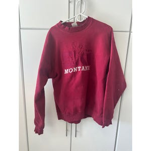 May include: A maroon sweatshirt with the word "MONTANA" in white block letters. Above the text is a white graphic of a state seal. The sweatshirt has long sleeves and a crew neck.