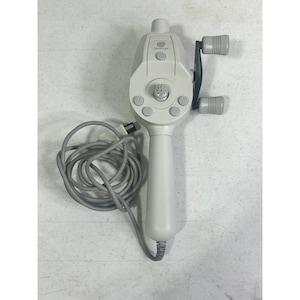 Può includere: Un controller a forma di canna da pesca Dreamcast grigio chiaro. Il controller ha una maniglia a mulinello, diversi pulsanti e un lungo cavo grigio. Il logo Dreamcast è visibile sulla parte superiore del controller.