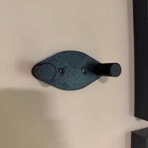 May include: A black wall-mounted coat hook with a unique, curved design. The hook features two cylindrical pegs and a central mounting plate, secured to the wall with screws. The hook is made of a textured material.