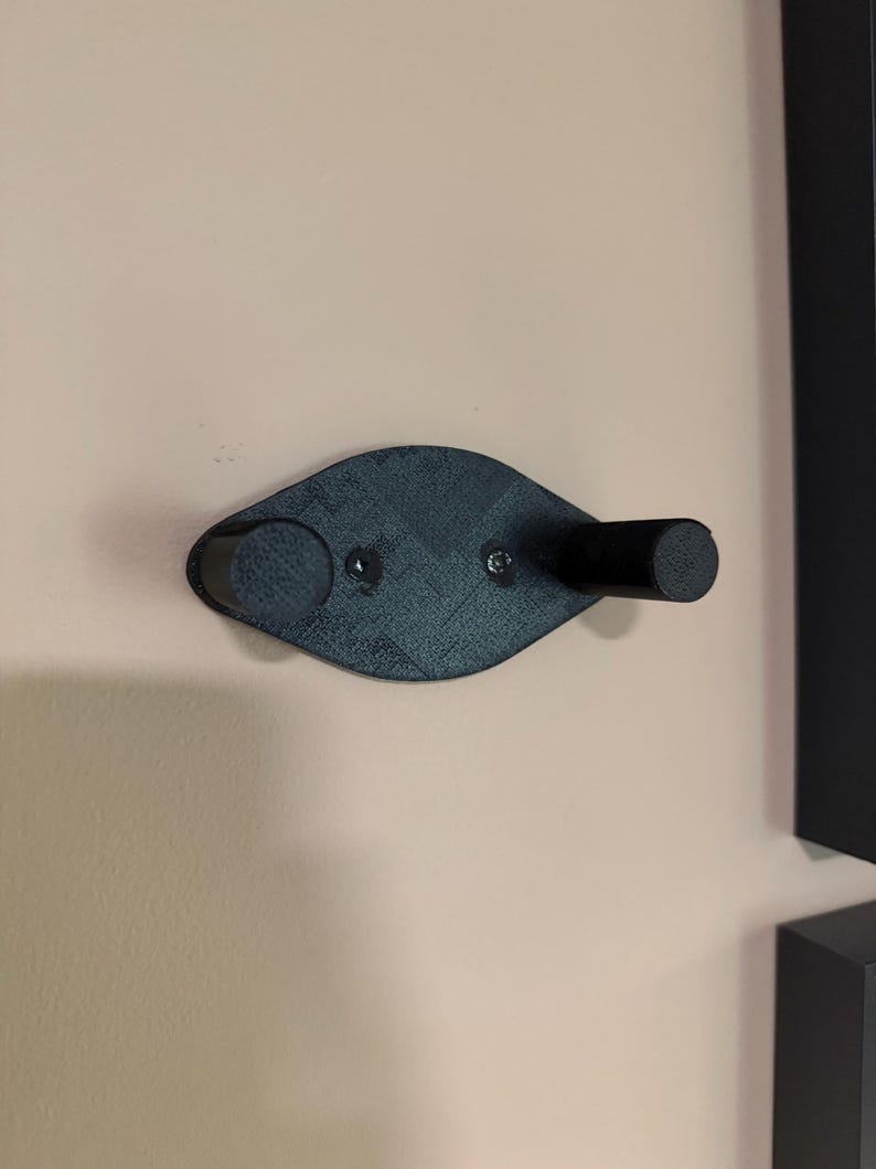 May include: A black wall-mounted coat hook with a unique, curved design. The hook features two cylindrical pegs and a central mounting plate, secured to the wall with screws. The hook is made of a textured material.