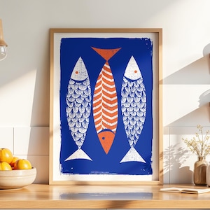 May include: A framed art print featuring three stylized fish against a blue background. Two fish are white with scale patterns, and one is orange with vertical stripes. The artwork is in a light wood frame.