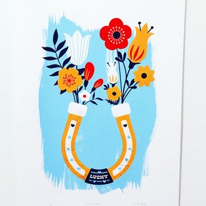May include: A yellow horseshoe with a blue banner that says "Lucky in Love" is filled with a bouquet of flowers. The flowers include red, yellow, white, and orange blooms. The background is a light blue color.