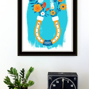 May include: A framed print of a colorful illustration of a horseshoe with flowers in the shape of a horseshoe. The horseshoe is orange and white, and the flowers are red, orange, yellow, white, and blue. The background is a light blue color with a white brushstroke effect. The text "LUCKY" is printed on the horseshoe.