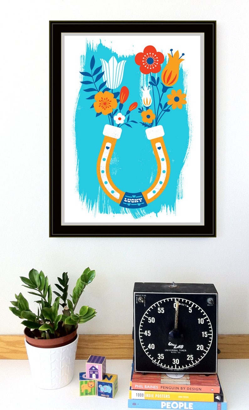 May include: A framed print of a colorful illustration of a horseshoe with flowers in the shape of a horseshoe. The horseshoe is orange and white, and the flowers are red, orange, yellow, white, and blue. The background is a light blue color with a white brushstroke effect. The text "LUCKY" is printed on the horseshoe.