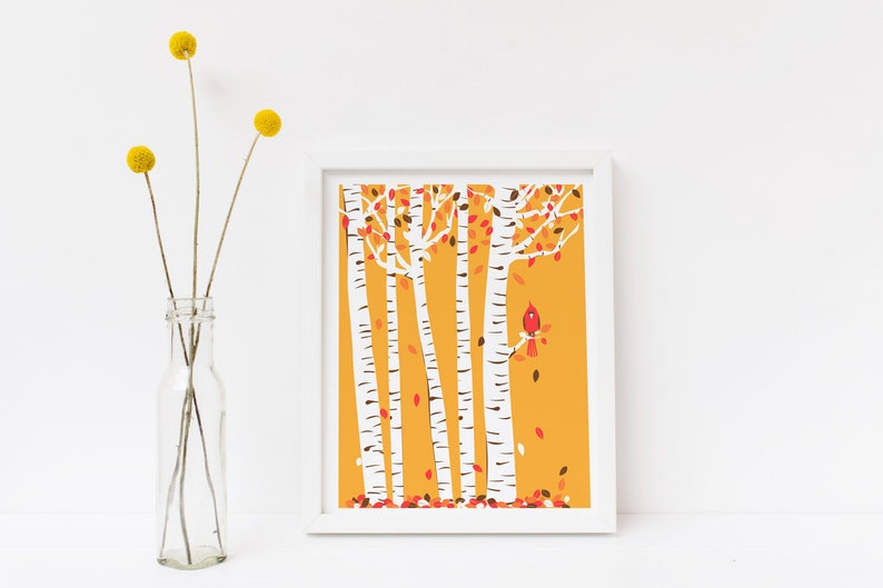 May include: A white framed print of a stylised illustration of a birch tree forest with a red cardinal perched on a branch. The background is a solid yellow colour with red and brown leaves falling from the trees.