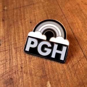 Pu&ograve; includere: Spilla in smalto nero e bianco con un arcobaleno e nuvole sopra il testo "PGH".