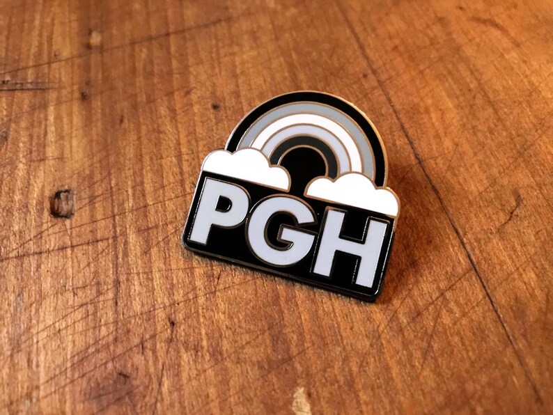 Pu&ograve; includere: Spilla in smalto nero e bianco con un arcobaleno e nuvole sopra il testo "PGH".