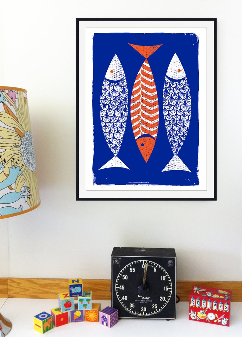 May include: A framed art print featuring three stylized fish against a blue background. Two fish are white with blue scale patterns, and the central fish is orange with a bone-like design. The print is displayed on a white wall.