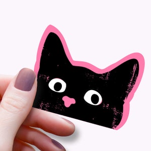 Könnte beinhalten: Ein schwarzer Katzenaufkleber mit rosa Umrandung. Die Katze hat große weiße Augen und eine rosa Nase. Der Aufkleber ist abgenutzt und hat eine strukturierte Optik.