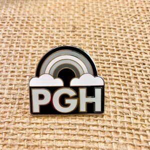 Pu&ograve; includere: Uno spillo in smalto bianco e nero con un arcobaleno e nuvole sopra il testo "PGH".