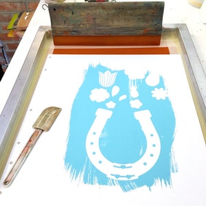 May include: A white horseshoe with a blue background and white flowers, leaves, and a tulip. The image is on a white surface with a wooden frame and a squeegee.