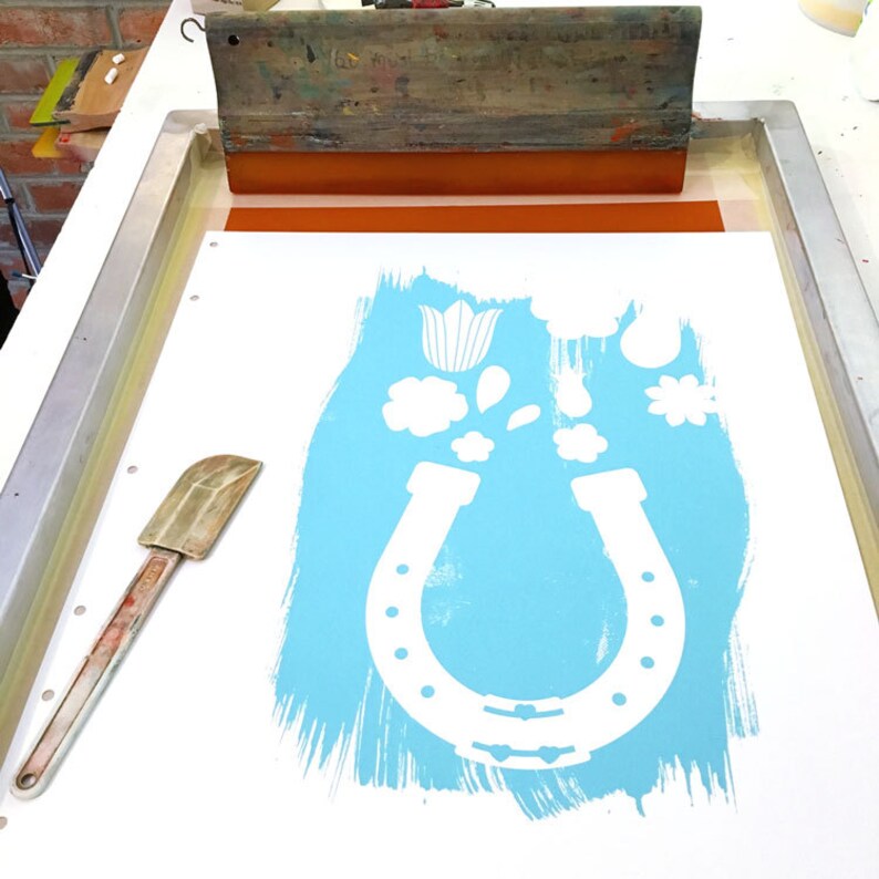 May include: A white horseshoe with a blue background and white flowers, leaves, and a tulip. The image is on a white surface with a wooden frame and a squeegee.