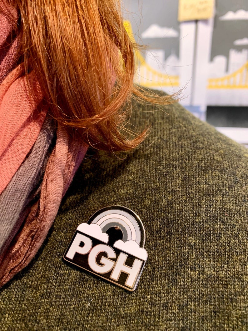 Pu&ograve; includere: Uno spillo in smalto bianco e nero con un arcobaleno e il testo "PGH" in lettere bianche. Lo spillo &egrave; su un maglione verde.