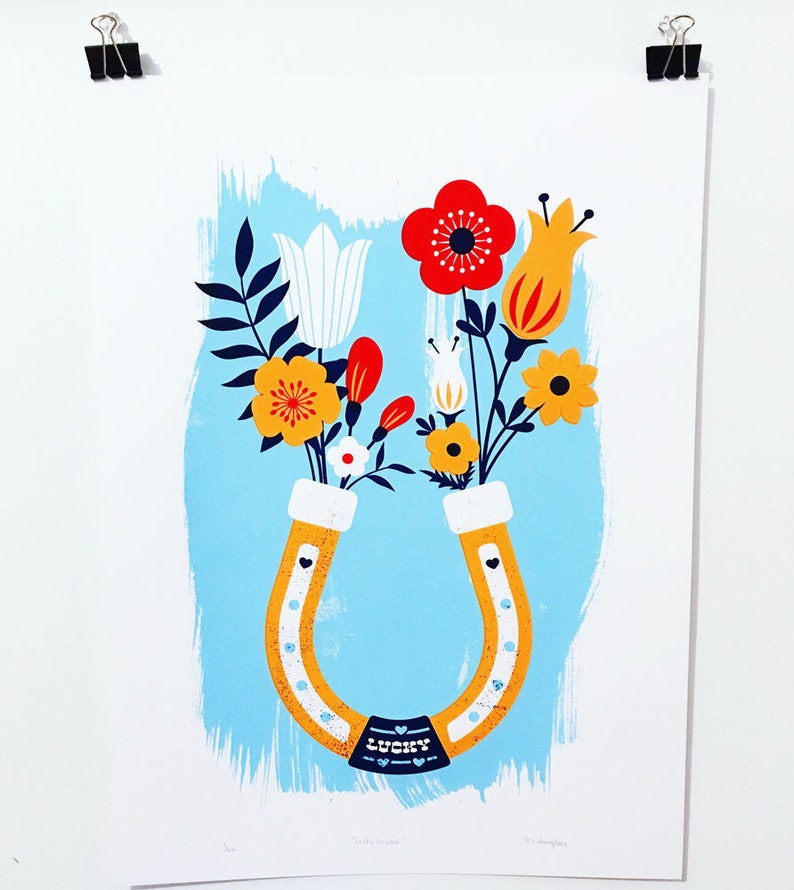May include: A yellow horseshoe with a blue banner that says "Lucky in Love" is filled with a bouquet of flowers. The flowers include red, yellow, white, and orange blooms. The background is a light blue color.