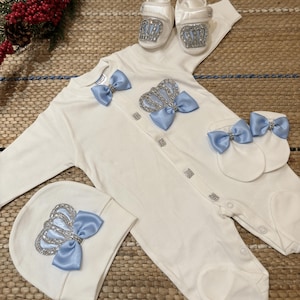 May include: A white baby onesie with a blue bow and a silver crown on the front. The onesie is part of a baby outfit set that includes a matching hat, booties, and a pair of mittens.