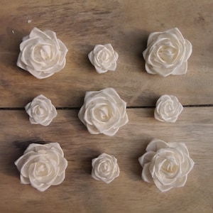 Puede incluir: Nueve rosas de madera blancas de diferentes tamaños dispuestas sobre una superficie de madera. Las rosas están todas mirando hacia la cámara y están enfocadas.