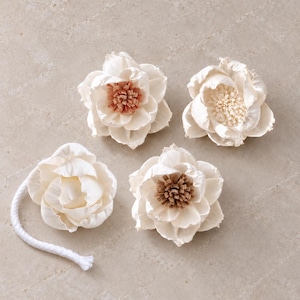 Op de afbeelding: Vier crèmekleurige decoratieve bloemen gemaakt van dun, gelaagd materiaal. Drie bloemen hebben opvallende centra in tinten lichtbruin, beige en crème. Eén bloem is voorzien van een lang, wit, gevlochten katoenen koord aan de onderzijde.