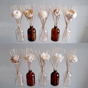 May include: Two rows of reed diffusers with decorative flower accents. Each diffuser features a clear or amber glass bottle filled with reeds and a decorative flower. The flowers are white, cream, and tan.