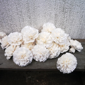 Może przedstawiać: Kolekcja białych sztucznych kwiatów goździków, ułożonych na zwietrzałej drewnianej powierzchni. Kwiaty mają wiele warstw delikatnych płatków, tworząc pełny, teksturowany wygląd. Tło to teksturowana, jasnoszara ściana.