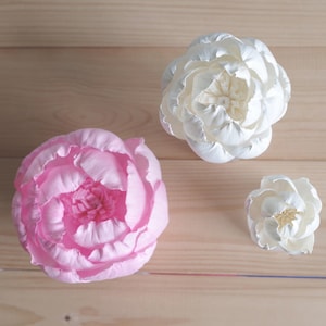 Puede incluir: Tres cabezas de flores artificiales, una rosa y dos blancas, sobre una superficie de madera. Las flores están hechas de tela y tienen un aspecto realista.