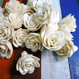 以下が含まれることがあります： カラフルな木製の背景に配置された白いソラウッドフラワーローズの集まり。バラは軽量で自然な素材で作られており、繊細でリアルな外観をしています。