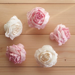 Puede incluir: Cinco rosas de papel artificiales en tonos de rosa y blanco dispuestas sobre una superficie de madera marrón claro. Las rosas están todas cerradas y tienen un aspecto delicado y en capas.