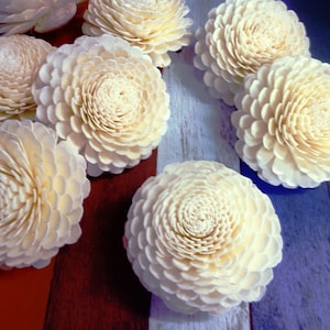 May include: A collection of eight white, intricately carved, wooden flower decorations. The flowers are arranged on a wooden surface with a blue and white painted design.