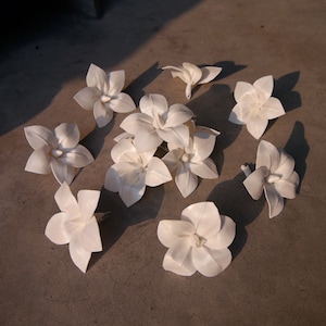 Può includere: Una collezione di dieci fiori di carta bianchi, ognuno con cinque petali e un punto centrale. I fiori sono disposti su una superficie marrone chiaro.