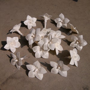 Op de afbeelding: Een verzameling van 15 witte papieren bloemenversieringen met draadstengels. De bloemen zijn in een cirkel op een grijs oppervlak gerangschikt.