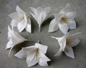 May include: Six white paper lily flowers with gold stamens. The flowers are arranged in a circle on a grey surface.