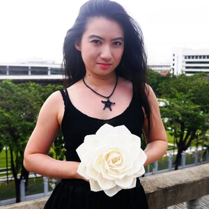 Könnte beinhalten: Eine Frau in einem schwarzen Tanktop mit einer schwarzen Sternen-Halskette hält eine große weiße Papierblume.