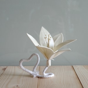 May include: A white paper flower with a rope heart wrapped around the stem. The flower is sitting on a light wood surface.