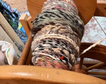 May include: A wooden spinning wheel bobbin filled with thick, hand-spun art yarn. The yarn features a variegated mix of colors including muted greens, browns, creams, and hints of orange, with varied textures and thicknesses wound around the central spool.