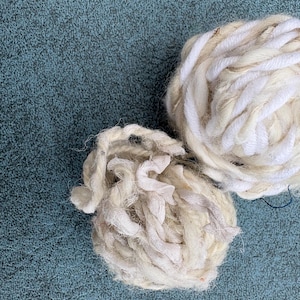 May include: Two balls of yarn in shades of off-white and cream. The larger ball features a thick, twisted white fibre, while the smaller ball is composed of a mix of textured, frayed, and ribbon-like off-white strands wound into a loose sphere.