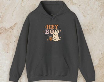 May include: A dark charcoal gray hooded sweatshirt featuring a graphic print on the chest. The design includes the text HEY BOO in orange and pink, a small white ghost holding an orange pumpkin mug, and several small black bats and stars.