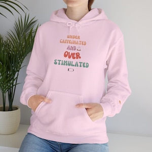 May include: A pale pink hoodie with a front pocket and a drawstring hood. The text "UNDER CAFFEINATED AND OVER STIMULATED" is printed on the front in various colours. The hoodie is made of a soft material.