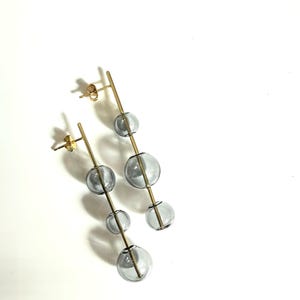 May include: A pair of gold-colored earrings with a vertical bar design. Each earring features three clear, gray-tinted glass spheres of varying sizes. The earrings have a post-back closure and are set against a white background.