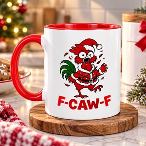 Op de afbeelding: Een witte keramische mok met een felrode binnenkant en handvat. De voorzijde toont een cartoonhaan met een kerstmuts en kerstverlichting, met de tekst F-CAW-F in dikke rode letters onder de illustratie gedrukt.