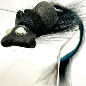 May include: A black and white fly fishing lure with a fuzzy tail and two white eyes. The lure is displayed on a clear acrylic stand.