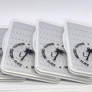 May include: Four gray fly fishing boxes with clear lids are stacked. Each box has a black graphic of a fly and hook, with the text "QUALITY - CUSTOM - FLIES" and "flyenthusiast.com" printed on the lid.