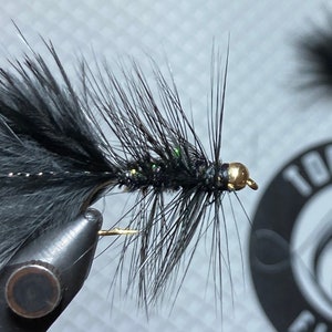 Puede incluir: Señuelo de pesca con mosca negro con detalles verdes y dorados. El señuelo está atado con plumas negras y tiene una cabeza de cuentas doradas.