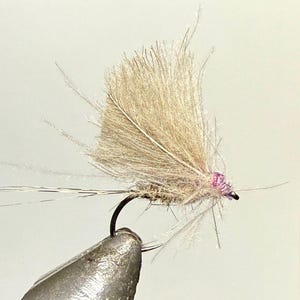May include: A close-up of a handmade fishing fly. The fly features a tan feather wing and a purple body. The hook is black and attached to a silver metal clamp.