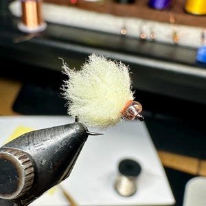 Può includere: Primo piano di un materiale da legatura di mosche di colore giallo chiaro con una testa di perline in rame. Il materiale è tenuto da una pinza.