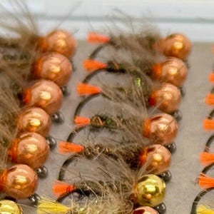 Può includere: Primo piano di un vassoio di esche per la pesca a mosca. Le esche sono legate con piume marroni e verdi e hanno perline di rame. Le esche sono disposte in file su un vassoio bianco.