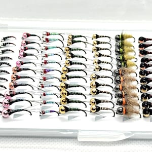 May include: A white plastic box filled with various fishing flies. The flies have black, gold, and silver hooks with colorful beads and feather accents. Some flies have a black and white striped pattern.