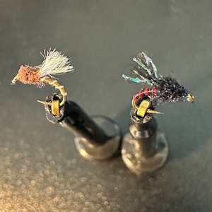 May include: Two fly fishing lures on black display stands. The lure on the left is brown with white feathers and a gold hook. The lure on the right is black with iridescent feathers and a red and gold hook.