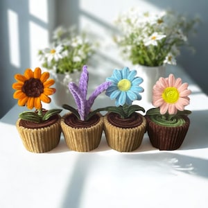 May include: Four decorative cupcakes designed to look like potted flowers. Each cupcake has a different flower: an orange sunflower, a purple flower, a blue daisy, and a pink daisy. The cupcakes are in brown paper cups, with green frosting leaves.